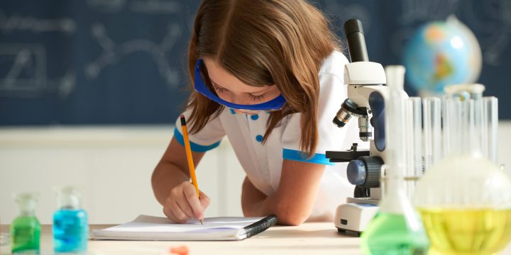 Little girl in goggles writing in textbook at chemistry class